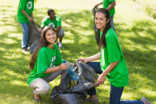 Four volunteers wearing bright green T-shirts are engaged in a community clean-up on a grassy outdoor area. In the foreground, two young women with long dark hair are kneeling on the grass, actively participating in rubbish collection; one is holding a large black garbage bag open while the other is placing a bottle into it. Behind them, two more volunteers, also dressed in green T-shirts, are standing and holding a separate black rubbish bag, contributing to the collection effort. The background features a lush, sunlit green space with scattered trees and shaded areas, indicating a park or communal outdoor setting. The volunteers are focused on gathering waste, emphasizing environmental responsibility. The scene captures the collaborative atmosphere typical of community litter-picking activities, aligned with rubbish removal initiatives in local areas such as Forest Hill, supporting environmental cleanliness efforts.